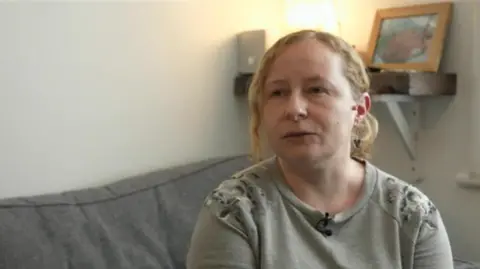 BBC Katie Fowler wears a grey jumper with a flower pattern on the shoulder. She has blonde hair which is mostly tied back with a curled part loose near the front. She is sitting on a grey sofa and in the background a photo of her baby is seen on a shelf.