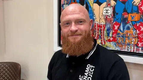 A man who is bald, wearing a black T-shirt and has a long ginger beard. He is smiling at the camera and sitting in front of a brightly coloured picture that is hanging on the wall.