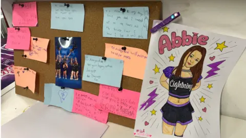 A noticeboard at Swindon Lightning Cheerleading Club covered in pink, orange and blue post-it notes with messages written in memory of Abbie, alongside a picture drawn of Abbie wearing the Lightning Cheerleading kit.