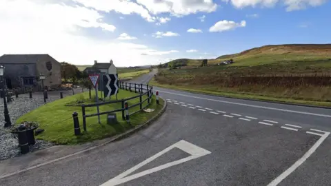 Google A Google street view of a rural road in the middle of fields and hills, viewed from a junction. On the left are several houses, a fence and road signs. 