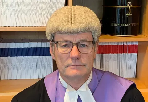 Judge Steven Parker, dressed in a wig and robe, looks into the camera in front of a bookshelf