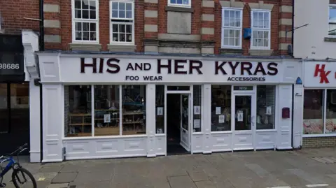 Front of the old His and Hers footwear store, some of the letters from the sign are missing, the storefront is painted white.