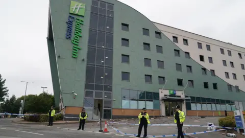 PA A wider shot of police officers at the Holiday Inn Express in Tamworth, Staffordshire. They are wearing uniform and stood in front of blue police tape. In the background we can see part of the building has been smashed up with rocks and glass on the floor. We can see the entire hotel in the background. 