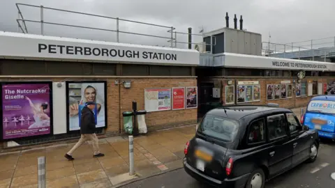 Street view image of Peterborough Train Station. The sky is a dark grey and there are several taxis parked outside the entrance. There are lots of advertisement boards on the brick exterior wall. 