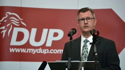 DUP leader Jeffrey Donaldson gives a keynote speech at the La Mon House Hotel on September 9, 2021 in Belfast, Northern Ireland.  He is in front of two microphones wearing a dark suit, white shirt and patterned tie. A red sign is behind him with the words DUP www.mydup.com in white.