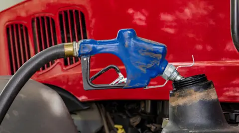 Getty Images Photograph of a blue diesel pump resting on the fuel cap of a red tractor. The pump and the fuel cap are slightly splashed with mud. 
