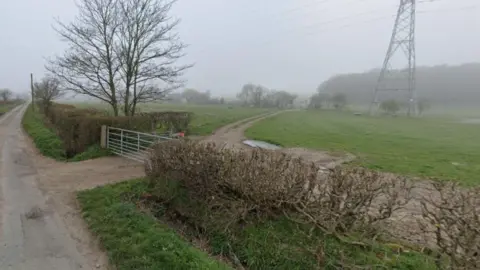 A road is on the left, with one exit off it leading to a gate. Behind the gate are fields of grass and in the distance, trees and an electricity pylon