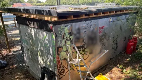 Charlie Stubbs / BBC There is multi-coloured graffiti on a grey metal wall. It includes big symbols and two blurred out images. There is also a metal ladder in front of it and two branches on the top. To the far right, there is two red gas cannisters