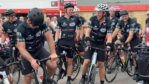 Image of a number of men in cycling gear, standing with their bicycles. In the background can be seen a crowd of people and the outside of a large stadium building