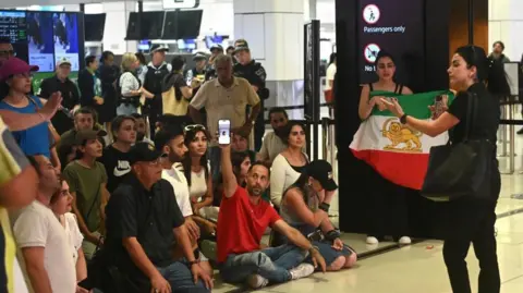A group of around 15 people, some seated, some standing, at the airport. Some with the Lion and Sun flag, which served as the official state flag before the Islamic revolution in Iran
