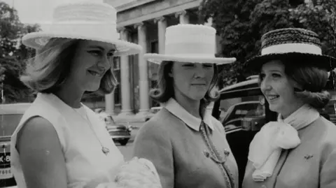 Shutterstock A black and white image from 1963 of three women stood together wearing wicker hats. A young Queen Camilla (L) wears a sleeveless dress with a pendant necklace next to two other women, one wearing a jacket with a collar poking out the top and the other wearing a neck scarf tied in a bow