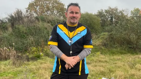Kevin Rudd, the acting secretary of Elmbridge Eagles Rugby Club. Kevin is standing at the Oaken Lane site and is wearing training gear for the Elmbridge Eagles, including a whistle and grey undershirt.
