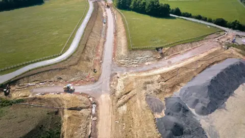 National Highways Another aerial picture shows a cross-shaped section of road dug out of green fields. A huge amount of sand-coloured cleared dirt is in piles beside the new route. A steamroller can be seen on the cleared track.