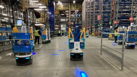 Robots in a warehouse - they are white and each carry two blue containers with stock in  