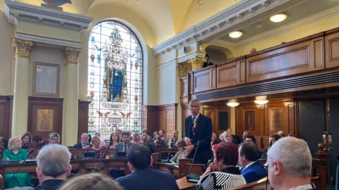 A man in a suit stands up speaking in an ornate council chamber full of elected members. There is a large stain glass window behind the councillors in Colchester City Council's main chamber.