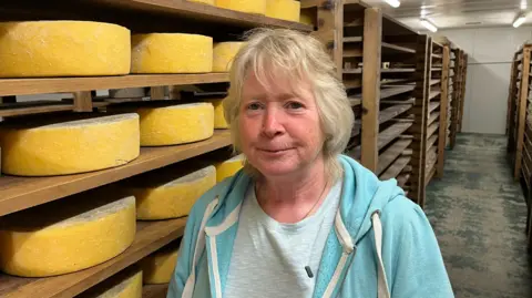 BBC/Phil Harrison Jane Bowyer, wearing a grey t-shirt and powder blue hoody, stands in front of stack of cheese wheels several feet high.