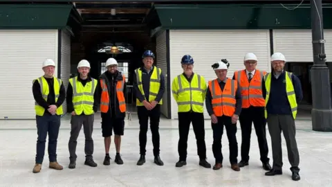 Pick Everard A group of people wearing yellow and orange high-visibility jackets are standing in a line, with hard hats on. They are in a market hall with stalls painted dark green with white shutters pulled down. There is an arched window in a recess behind the group.