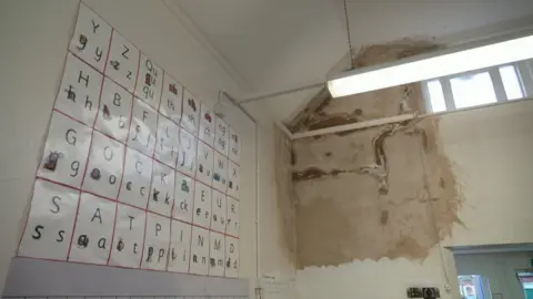 A classroom with an alphabet board on one wall. The wall next to it has a large, dirty, brown damp patch leading from the roof, where a leak has been