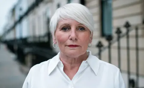 GMB Scotland A woman with white hair and large earrins, wearing a white blouse, looks directly at the camera