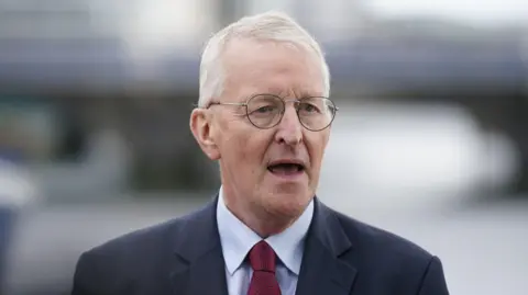 Headshot of Hilary Benn wearing a suit and tie. Background is out of focus.