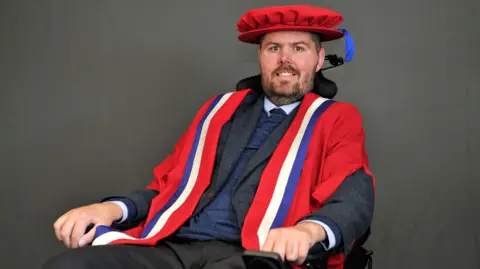 Ed Slater is sat in a wheelchair. He is smiling at the camera, dressed in graduation robes and cap. He is wearing a suit.