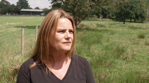 Joanna Drake from Protect Lindow has shoulder length dark blonde hair and is wearing a grey v-necked top. She is photographed in a grassy field, with trees behind her and a wire fence to one side.