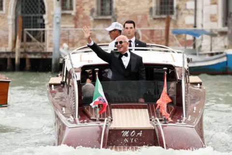 Getty Images Jeff Bezos in dark glasses and tuxedo waving from the back of a small motor boat in Venice on 27 June 2025.