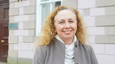Jersey deputy Inna Gardiner has a broad smile while standing outside a government building. She is wearing a grey goat, white turtle neck jumper and has long curly blonde hair.