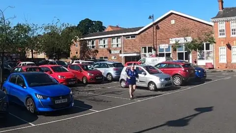 A woman walking across an empty parking bay in a car park, which has a dozen or more cars parked in front of a row of buildings