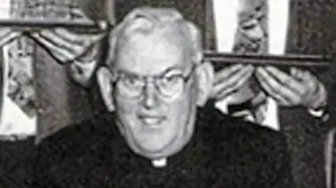 Pacemaker A black and white photo of the late Fr Malachy Finegan. He is wearing glasses, a black suit and a priest's collar.