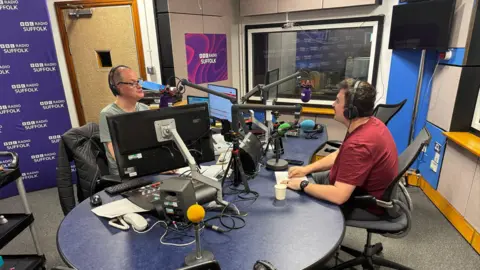 George King/BBC Two presenters sit at a desk speaking into radio mics. 