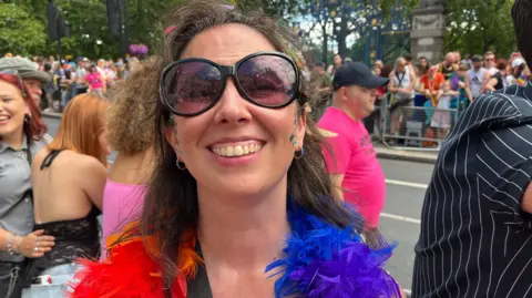 A close-up shot of Nicole Edmondson smiling at the camera, wearing large dark sunglasses and a vibrant red, orange, and blue feather boa amidst a busy outdoor crowd.
