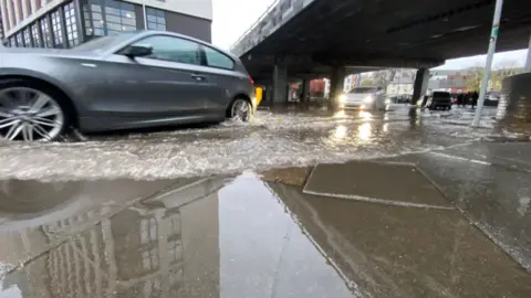 BBC A car drives through surface water in Coventry. Other vehicles are behind, driving under a bridge and there are buildings and houses in the background.