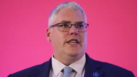 PA Media Gavin Robinson pictured in a suit in front of a pink background