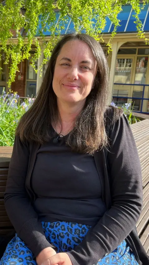 A woman wearing a black top and blue trousers is sitting outside under a tree.