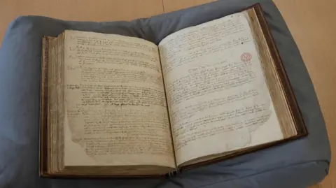 Qays Najm/BBC An open page of the Red Register on a wooden table. The book is rested on a grey cushion.