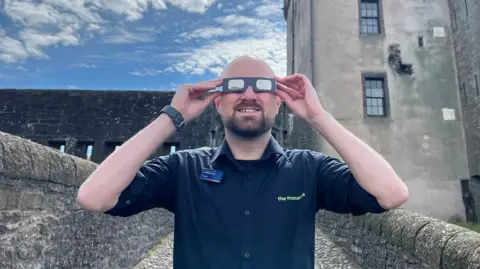 A man standing in front of Broughty Castle wearing eclipse glasses. He has on a dark blue shirt.