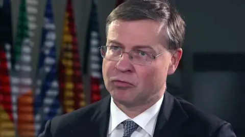 BBC Valdis is wearing a suit and a patterned tie. He is sitting in a room and behind him is a row of colourful national flags displayed in the background.