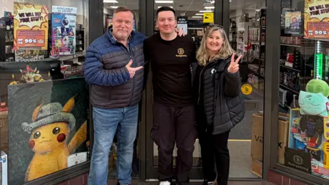 Alex Badham Shop owner Alex Badham outside his gaming shop, he's smiling and stood next to a man and a women. Alex has short dark hair, is cleanly shaven and is wearing a black t-shirt and trousers. The woman next to him is making a piece symbol with her hands and smiling, while the man on Alex's other side is pointing at Alex and smiling. 