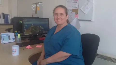 Nicole McKelvie has long brown hair tied back and is wearing blue scrubs and sitting at a desk with a computer monitor