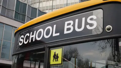The front of a bus, it is yellow and has the words school bus in white on the front. There is also a sign on the front depicting two children. There are buildings in the background of the bus.