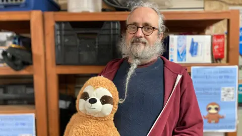 Nick Turner wearing a navy blue jumper underneath a red zip up hoodie. He has long grey hair tied back into a bun, and a long grey beard in a plait. He is wearing round, brown framed glasses and looking at the camera. In his right hand is a brown sloth stuffed toy. Behind him is a wooden shelving unit full of boxes and equipment.