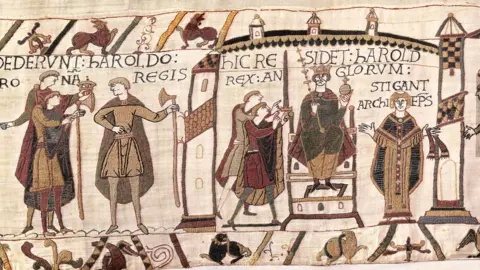 Bayeux Museum A wide crop of a scene involving Harold sitting on the throne holding an orb and stick along with figures to his left and right, some castle details, and other figures to the left of frame surrounde by birds and medieval lettering with fabric texture details.