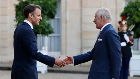 King Charles on the right of the picture holding out his right hand to President Macron, who is on the left of the picture, on the state visit to France in September 2023. Both men are wearing dark blue suits. 