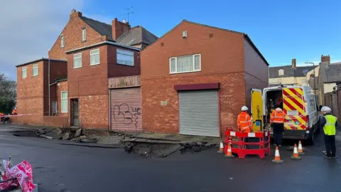 A collapsed road in front of red brick houses. Men in hi-vis are standing to the right.