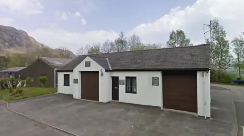 General view of a small building with parking space at the front. In the background is a Lake District fell.