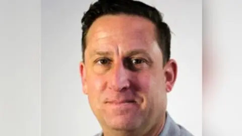 A headshot of a man wearing a red top and a grey jacket.