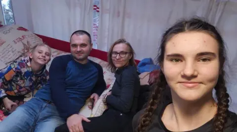 Family handout A girl with piggy-tail plaits with brown hair is stood up taking a selfie, with a man and woman and young boy sat on a bed next to her. They are all smiling, and the man's hand is resting on the woman's leg.