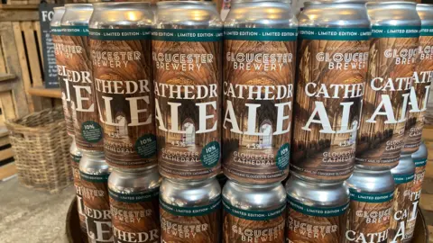 Stacked CathedrALE cans, metal cans of Gloucester Brewery, ale with an image of Gloucester Cathedral's cloisters, stacked in a room in the brewery, with wicker baskets and wood behind them