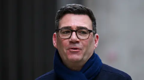Andy Burnham wearing a navy coat and dark blue scarf and black-rimmed glasses, looking to the side as he talks 